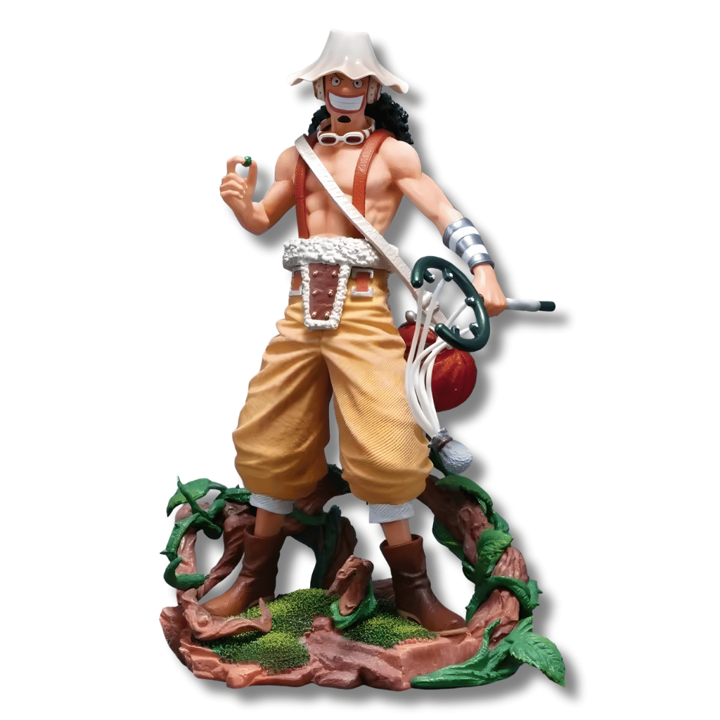 Una figura de estatua de Usopp del anime One Piece, de unos 25 cm de altura. El personaje está de pie sobre una base marrón con vegetación, vistiendo pantalones holgados de color canela y un arnés. Lleva su sombrero blanco y redondo, gafas y tiene su tirachinas grande colgado a un lado.
