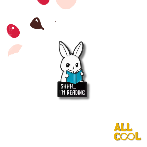 Un pin de metal esmaltado con el diseño de un conejo blanco sentado leyendo un libro. El conejo lleva un cartel que dice "Shhh... estoy leyendo" y tiene una expresión concentrada.