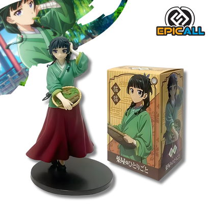 Una figura de estatua de Maomao del anime Diarios De La Boticaria. La figura viste una túnica verde y una falda roja larga, tiene el cabello oscuro recogido y sostiene una pequeña cesta tejida llena de verduras o hierbas con su mano derecha. Está de pie sobre una base circular negra.