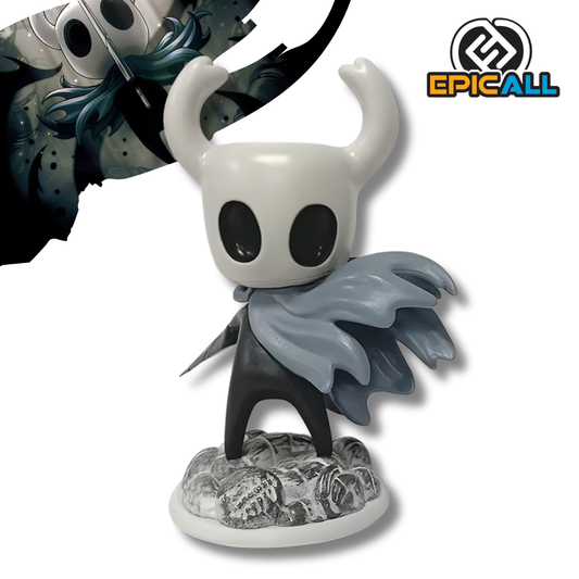 Una figura de estatua del personaje de videojuego Hollow Knight (el Caballerito), de unos 15 cm de altura. El personaje es negro con una máscara blanca con cuernos. Viste una capa gris que fluye dramáticamente. Está de pie sobre una base que simula rocas o nieve.