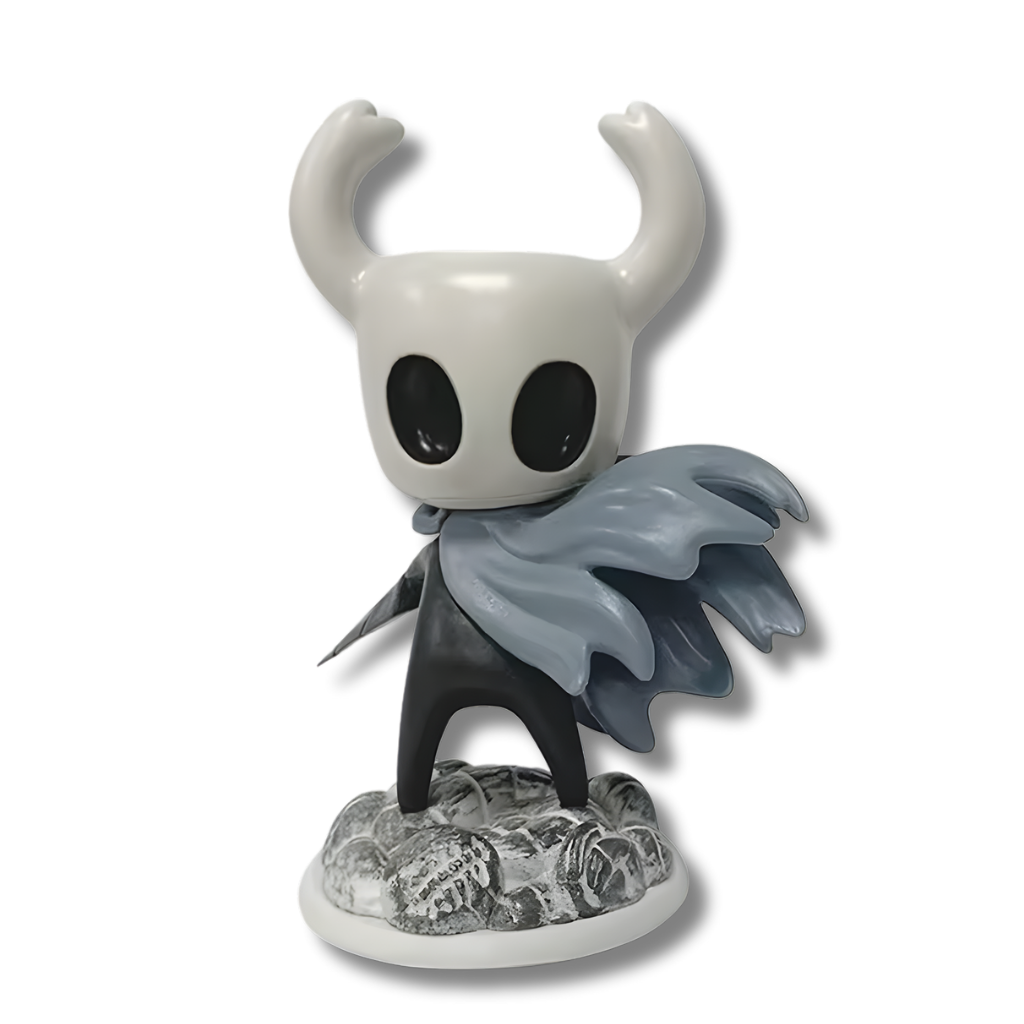 Una figura de estatua del personaje de videojuego Hollow Knight (el Caballerito), de unos 15 cm de altura. El personaje es negro con una máscara blanca con cuernos. Viste una capa gris que fluye dramáticamente. Está de pie sobre una base que simula rocas o nieve.