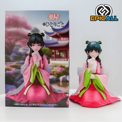 Figura de estatua de Maomao del anime Los Diarios de la Boticaria. El personaje aparece sentado con las piernas cruzadas ocultas por su kimono. Viste un traje tradicional compuesto por una túnica rosa sobre una falda roja, con amplias mangas de color verde brillante y un chal blanco translúcido. Maomao tiene el cabello oscuro recogido en dos moños laterales y sostiene un pequeño pañuelo blanco con su mano derecha cerca de su barbilla.