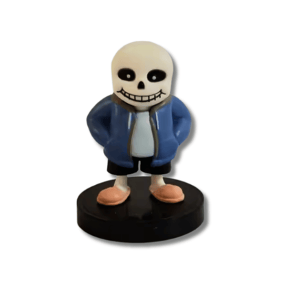 Una pequeña figura de estatua de Sans, el esqueleto del videojuego Undertale, de unos 7 cm de altura. El personaje tiene una cabeza de calavera blanca, ojos negros, viste una chaqueta azul con capucha, pantalones negros y pantuflas rosas, y está de pie sobre una base circular negra.