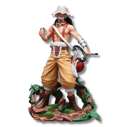 Una figura de estatua de Usopp del anime One Piece, de unos 25 cm de altura. El personaje está de pie sobre una base marrón con vegetación, vistiendo pantalones holgados de color canela y un arnés. Lleva su sombrero blanco y redondo, gafas y tiene su tirachinas grande colgado a un lado.