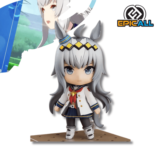 Figura pequeña de estilo Nendoroid del personaje Oguri Cap de Uma Musume Pretty Derby. Tiene cabello largo gris plateado, orejas de caballo y una diadema con rombos amarillos. Viste un uniforme escolar blanco con cuello de marinero azul y una falda azul marino. La figura está de pie sobre una base cuadrada con agujeros de color marrón claro.