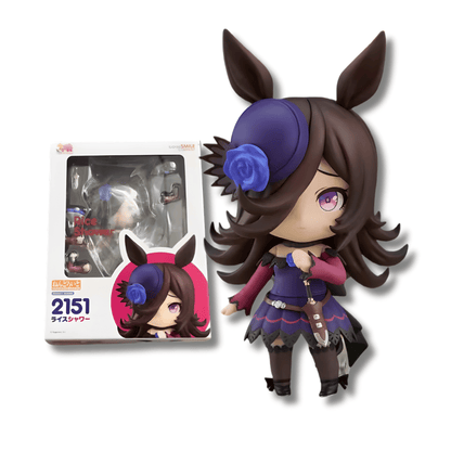 Figura de estilo Nendoroid del personaje Rice Shower de Uma Musume Pretty Derby. Tiene cabello largo castaño oscuro con orejas de caballo sobresaliendo y un pequeño sombrero morado con una rosa azul. Viste un traje morado con detalles rojos y un pequeño puñal en la cintura. Al lado izquierdo se observa su caja original de color blanco y azul con el número de serie 2151.