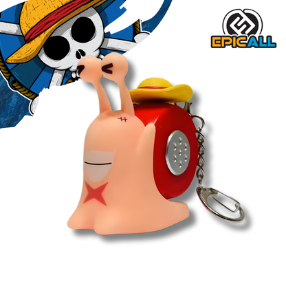 Un llavero con una figura de un Den Den Mushi (caracol telefónico) del anime One Piece. La figura es de color rosa pálido, tiene un caparazón rojo con detalles plateados y lleva un pequeño sombrero de paja amarillo y rojo. La figura tiene una cicatriz en forma de X en su mejilla izquierda y un aro de metal para llaves.