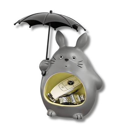 Un organizador con la forma del personaje de Totoro de color gris, sosteniendo un paraguas negro. El abdomen de Totoro tiene una abertura circular de color amarillo claro, donde se pueden colocar llaves, un reloj y un llavero de auto, visibles en la imagen.